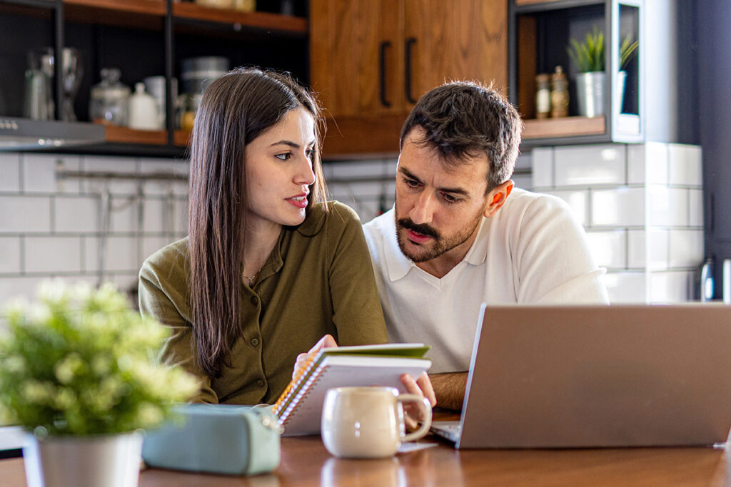Young couple calculating finances using laptop and notebook, managing their budget and expenses in their modern kitchen