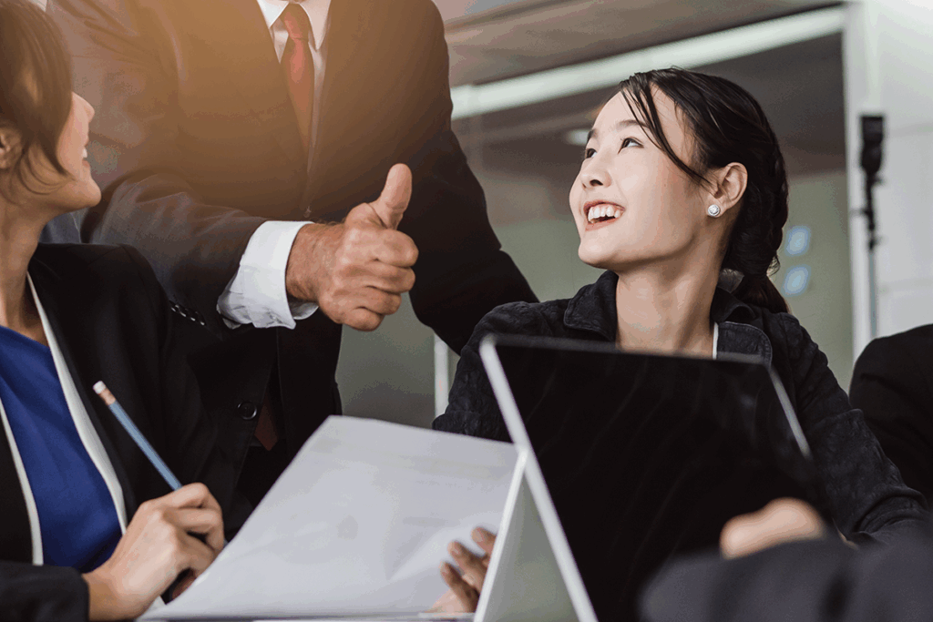 Positive work environment with boss in a suit giving the thumbs up to praise a smiling Japanese employee.