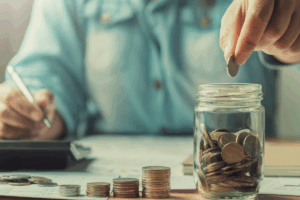 Person putting coins into a glass jar while calculating savings