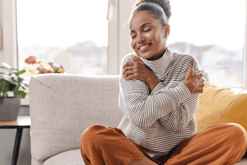 Woman sitting on a couch hugging herself with a smile, representing self-acceptance and emotional grounding.